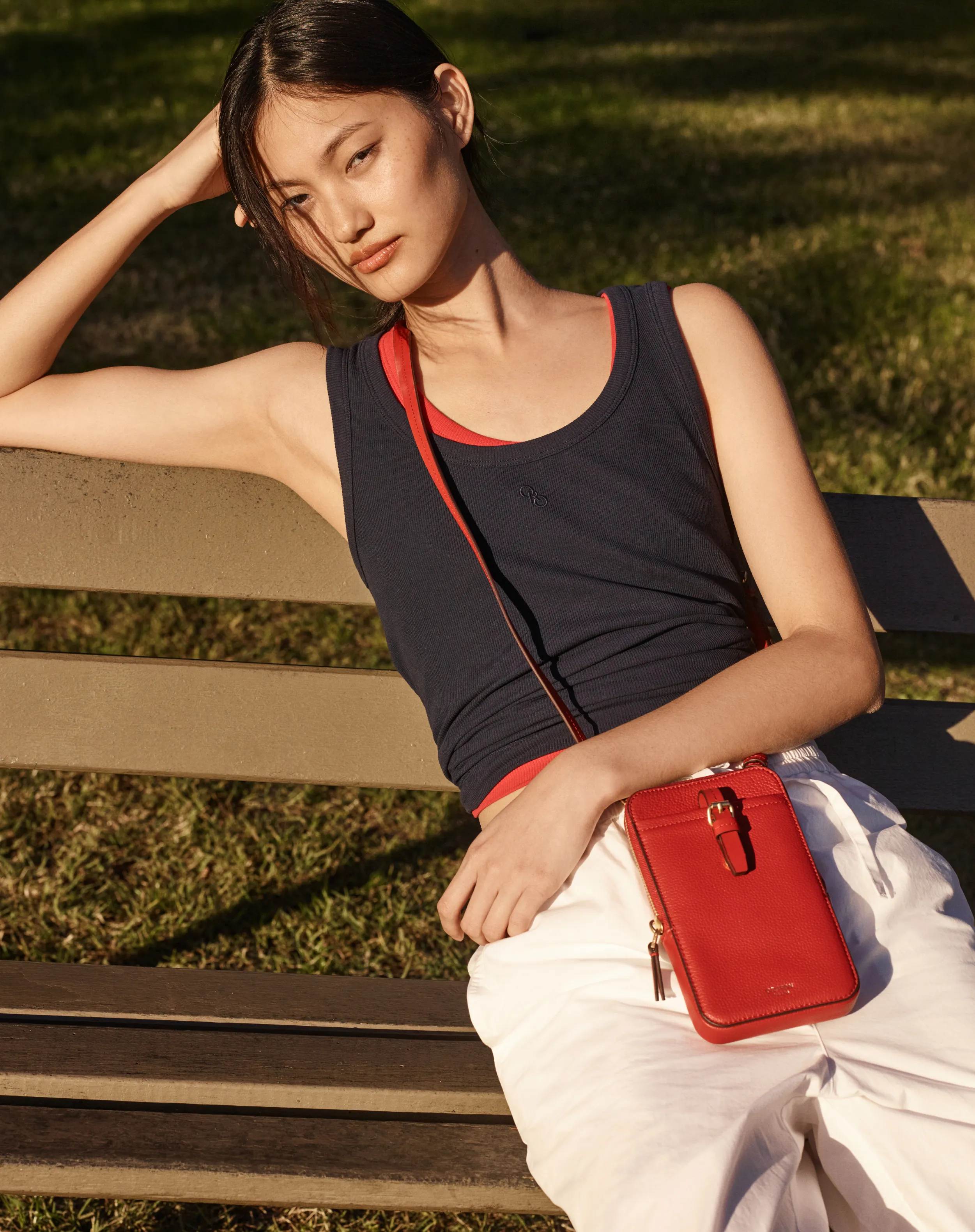 Young woman sitting on a bench, wearing a tank top and holding a red purse.