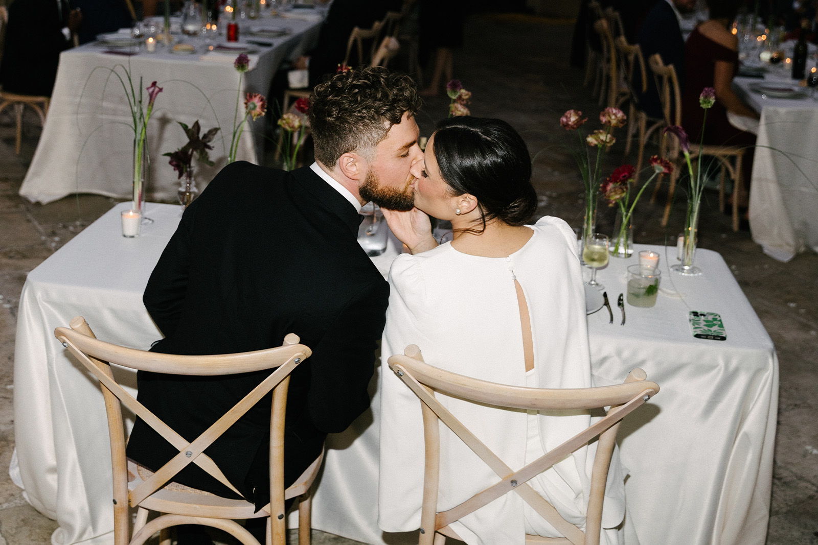 A couple kissing at a wedding reception, seated at a beautifully decorated table.