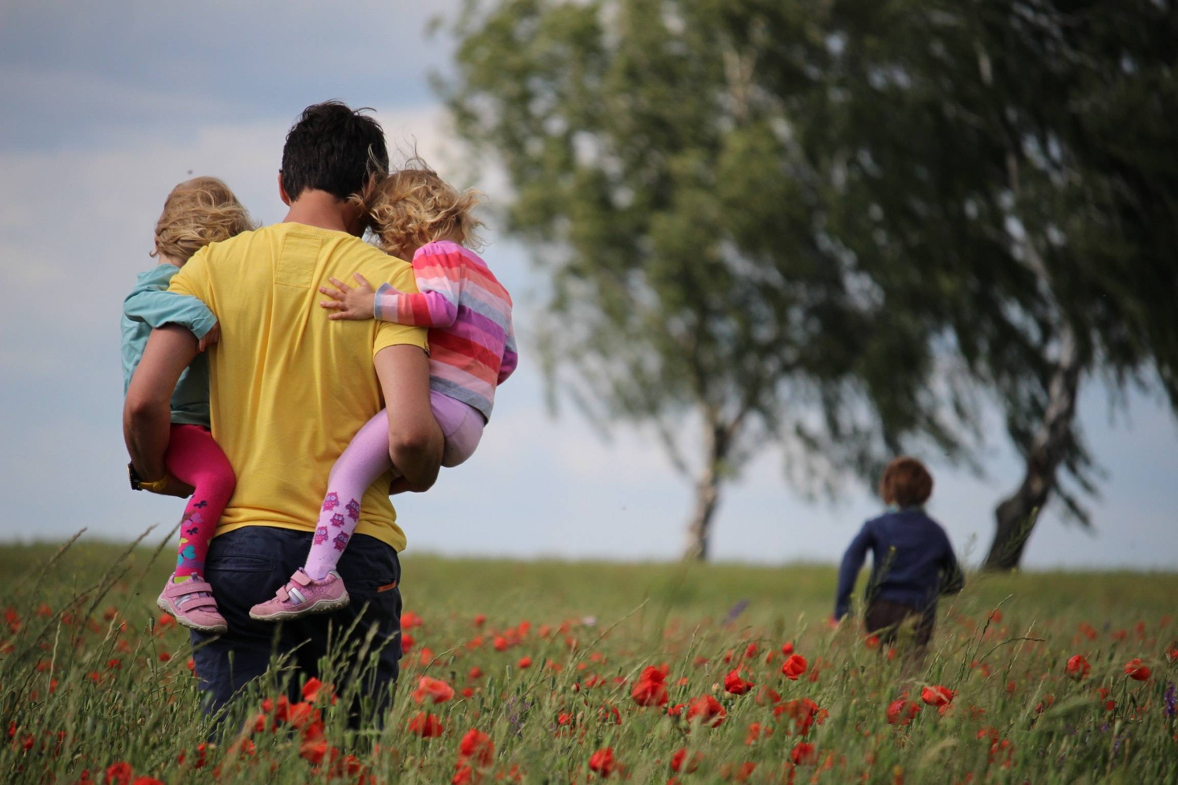 En far bærer to små jenter gjennom en eng med langt gress og valmuer, mens broren deres løper foran dem