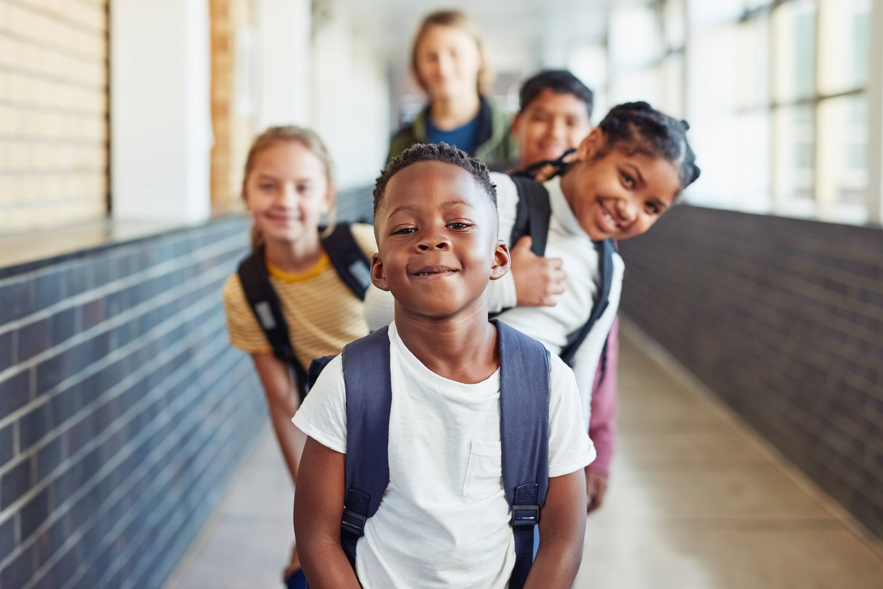 Kinderen staan in de rij in een schoolgang met hun leraar – achter het eerste kind steken een paar lachende kinderen links en rechts uit 