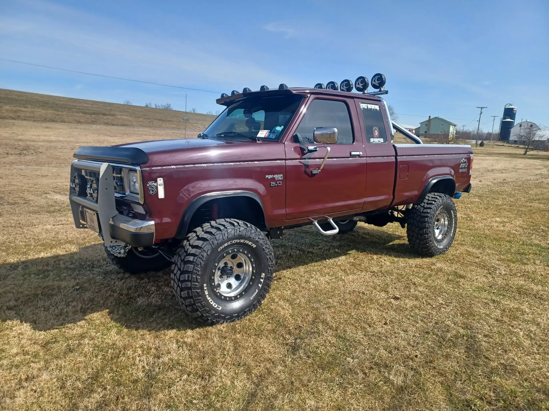 Front three-quarter Brenda's lifted maroon 1988 Ford Ranger with roll bar KC lights and Cooper tires in grassy field photo 1