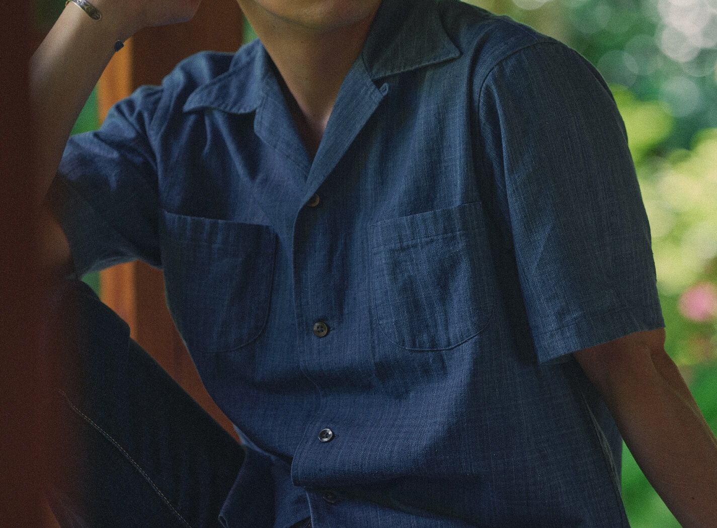 Person wearing a blue shirt, sitting outdoors with greenery in the background.