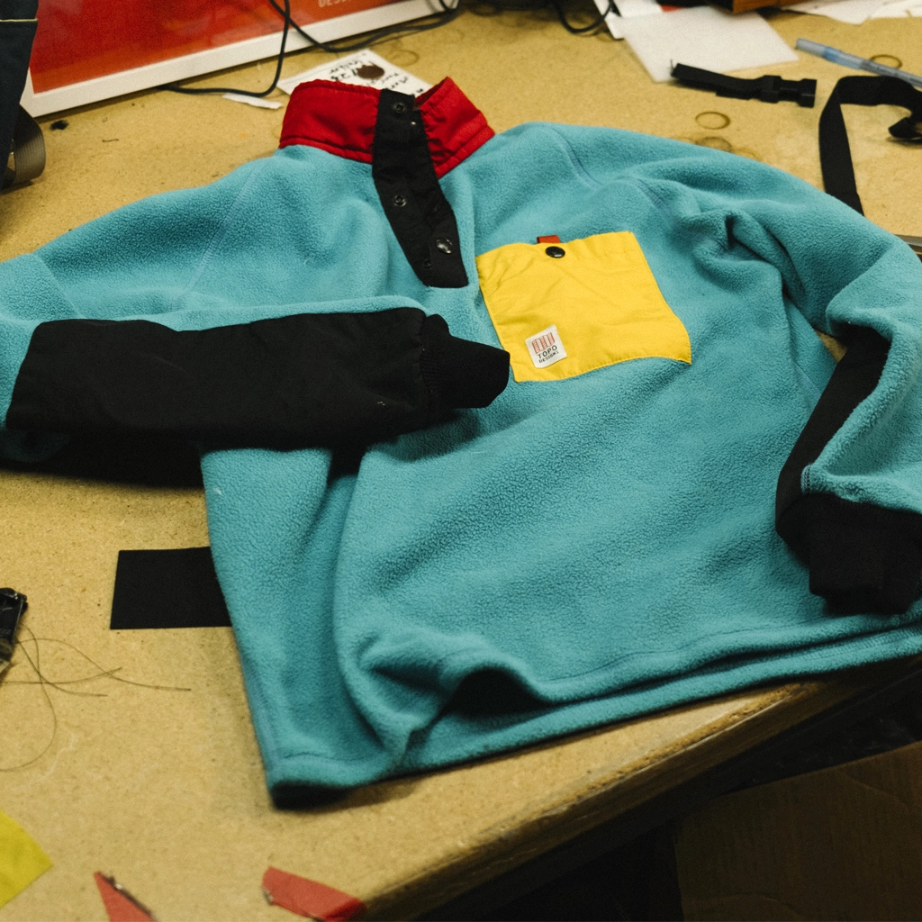 A turquoise fleece pullover with a yellow pocket on a worktable.