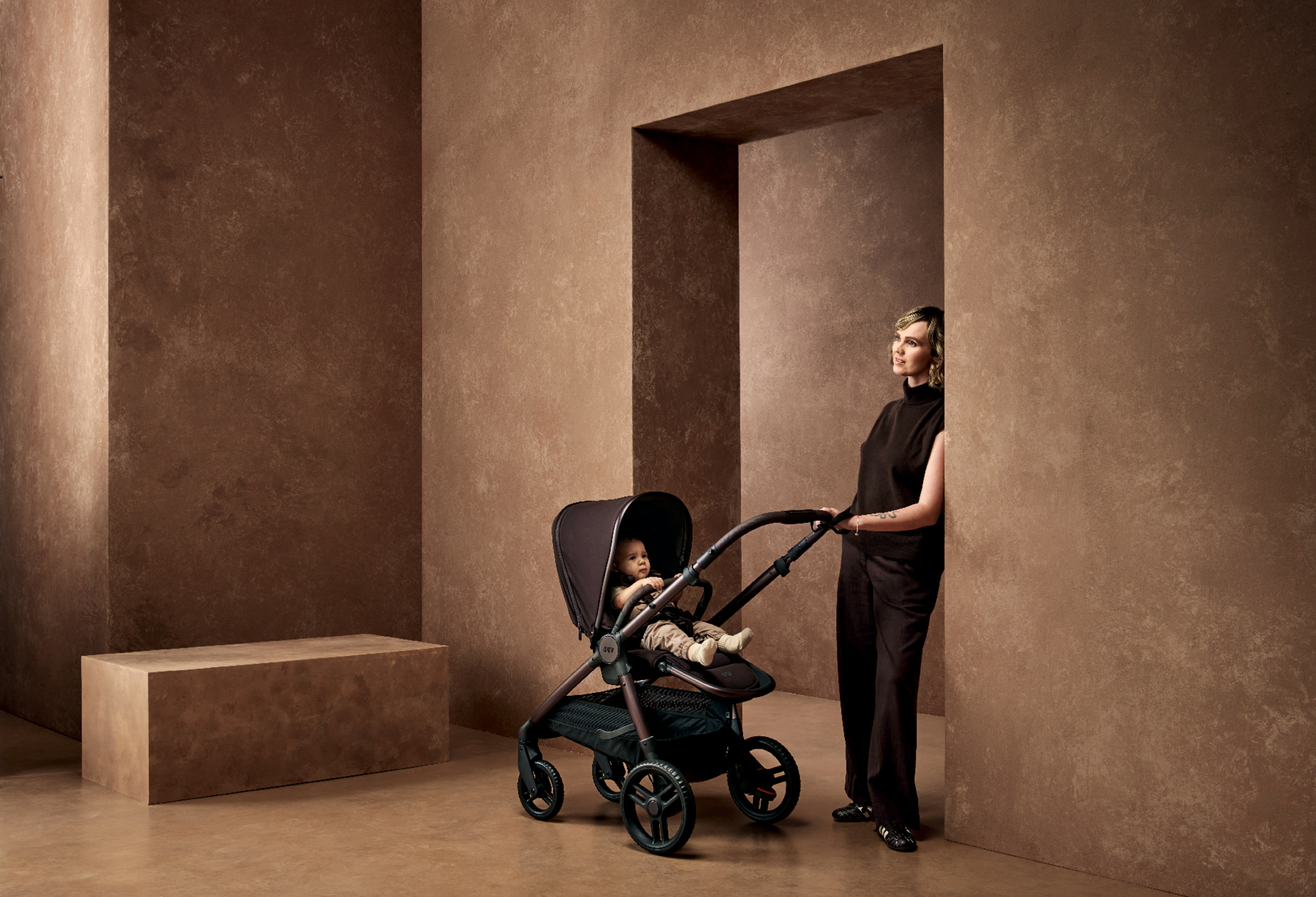 Person standing beside a stroller in a minimalist, earthy-toned environment.