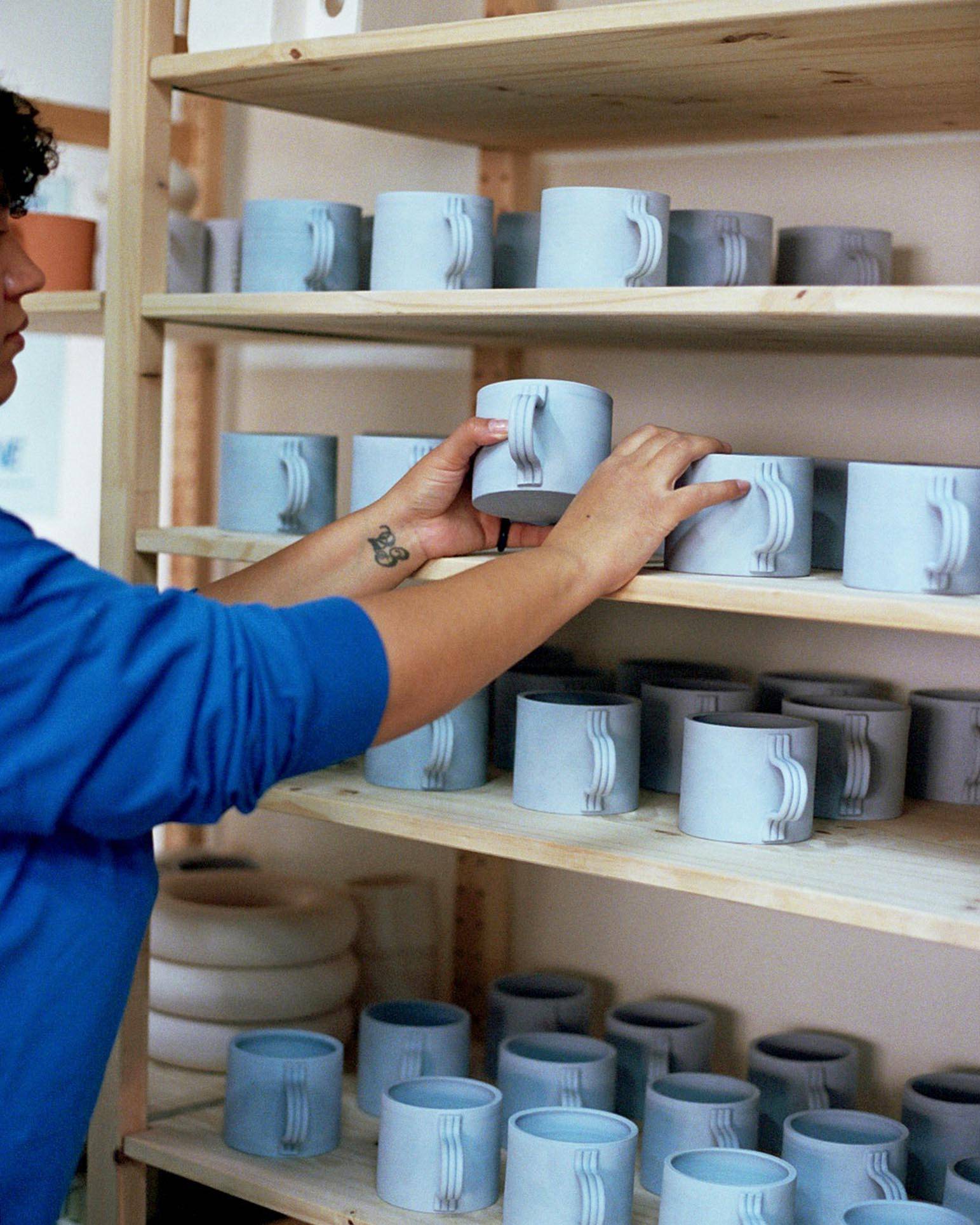 Detail image of Sara placing incomplete ceramic mugs on a shelf.