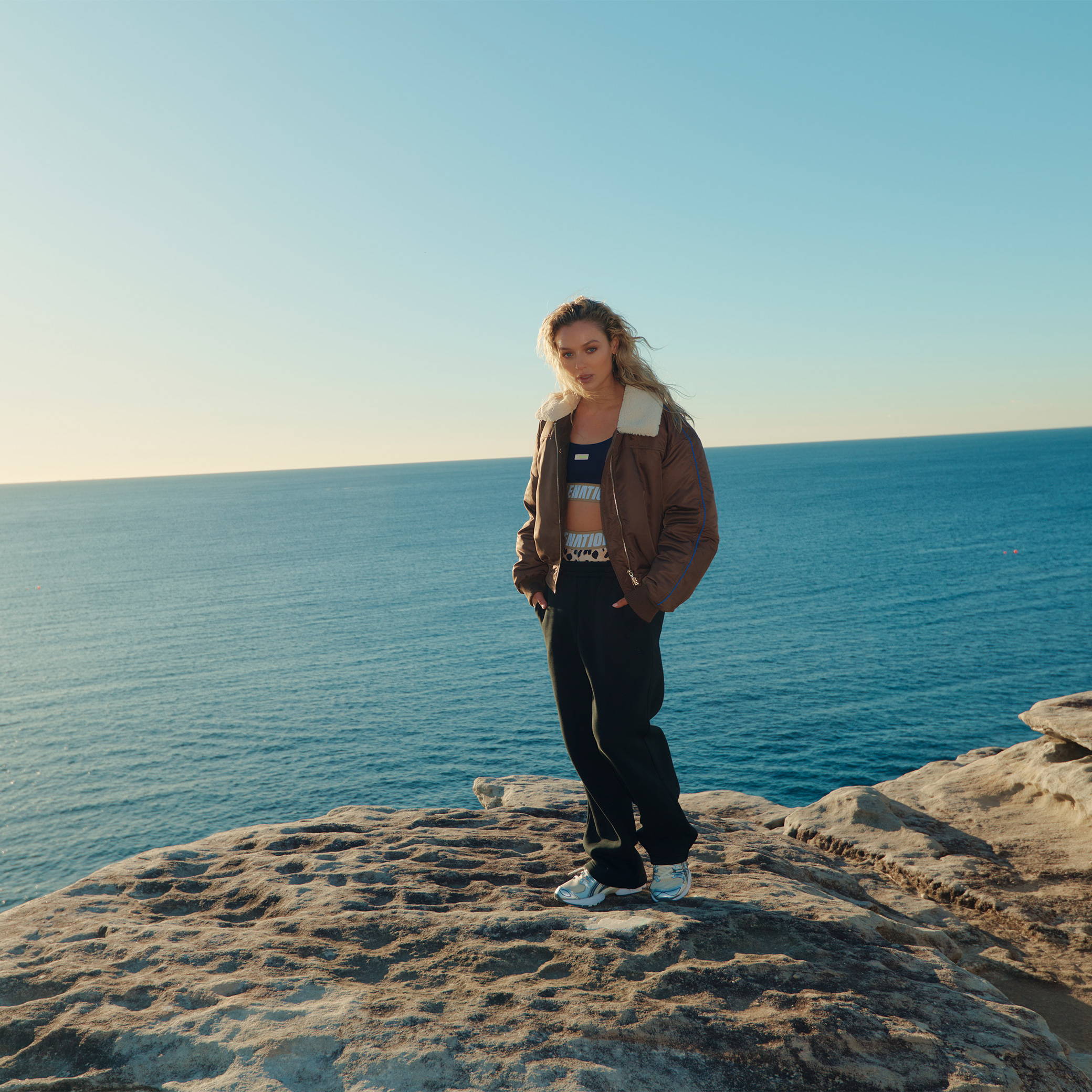 A girl is standing on a sandstone clifftop next to the ocean. She is wearing a warm jacket over trackpants and activewear. 