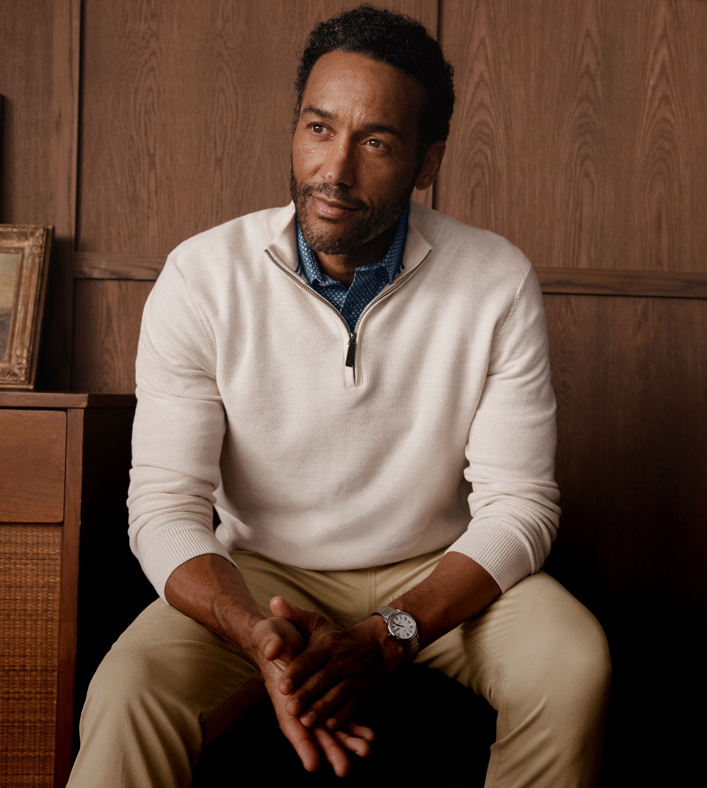 Man in a sweater sitting against a wooden background, looking thoughtfully.
