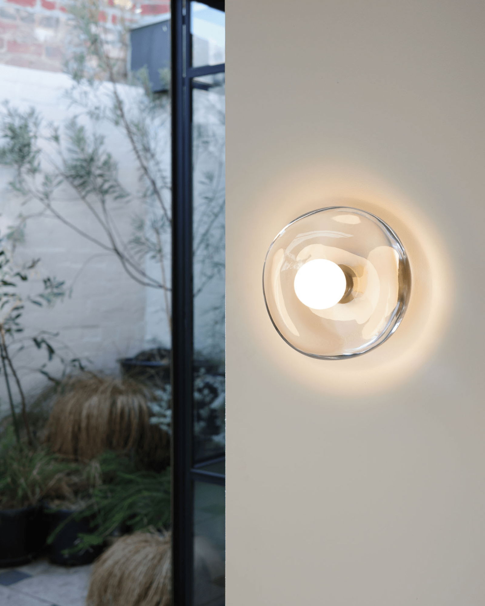 Handmade glass wall sconce on a white wall with a garden in the background light on