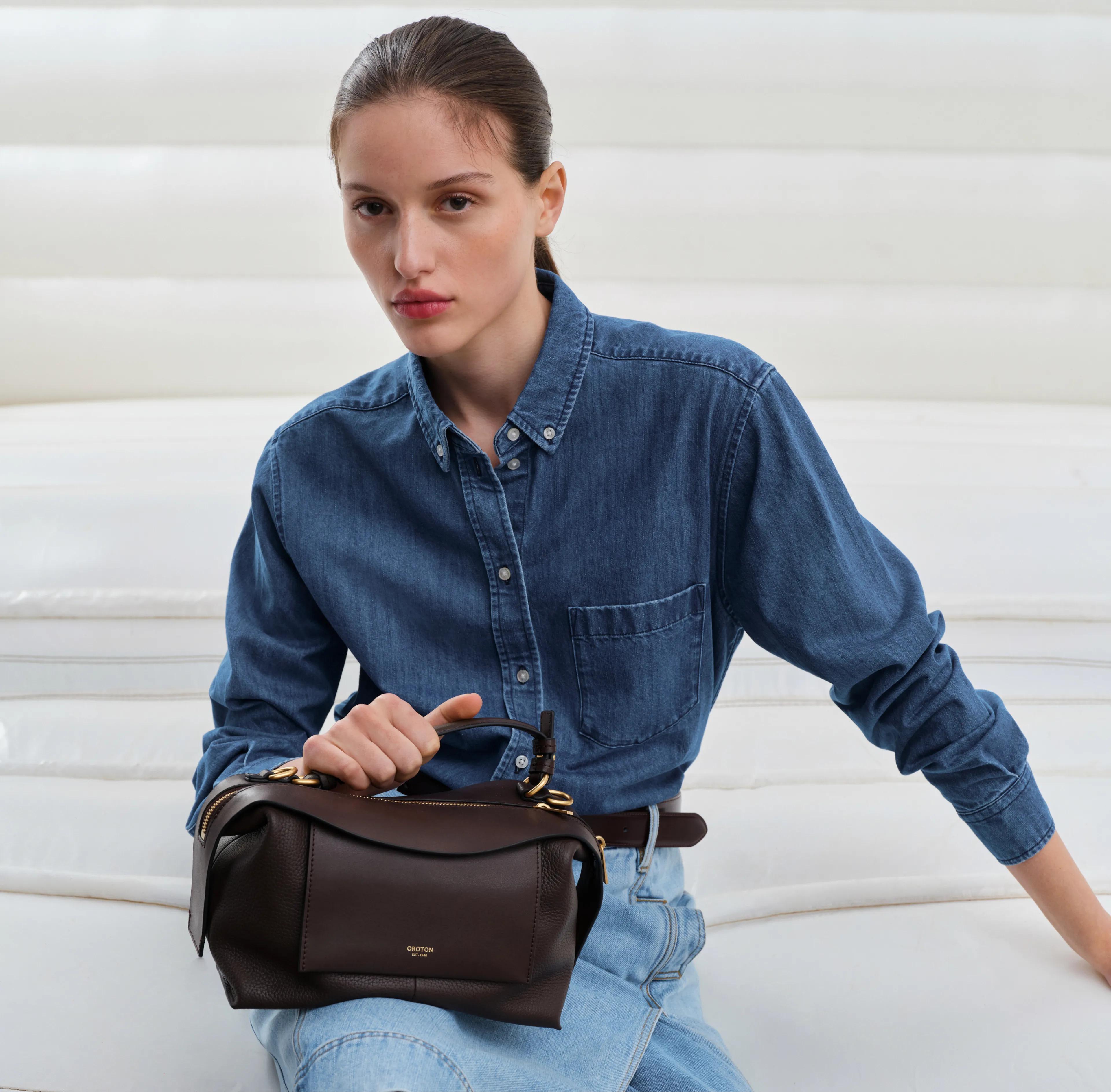 Model in denim shirt holding a brown handbag, seated on a white couch.