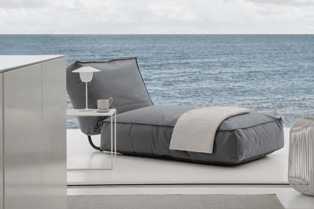 Low outdoor lounge daybed with gray cushions and a light throw beside a small side table, facing an ocean view.