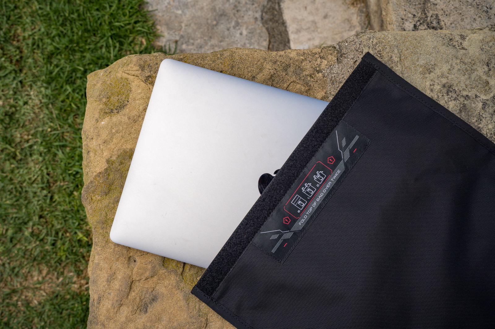 The bag outdoors with a laptop half inserted, showing the full-width Velcro strip and the printed closure diagram