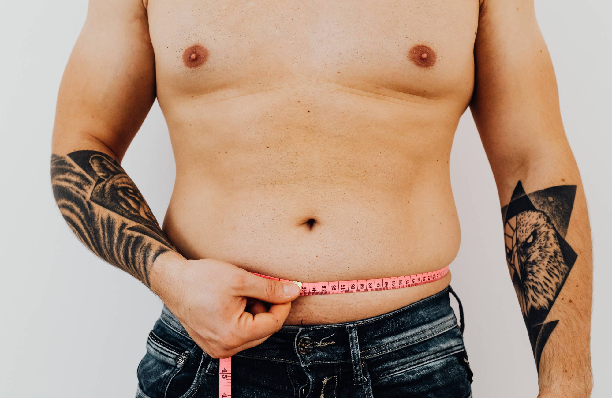  A shirtless man with arm tattoos wearing dark jeans measures his abdomen with a pink tape measurer.