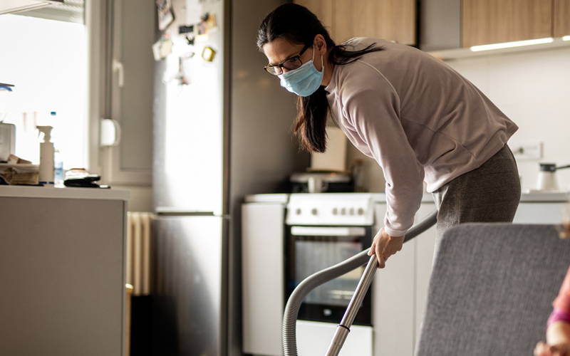 Žena s rouškou na tváři vysává v kuchyni. Pomocí roušky se snaží předejít vdechování alergenů, které se při vysávání rozvíří. 