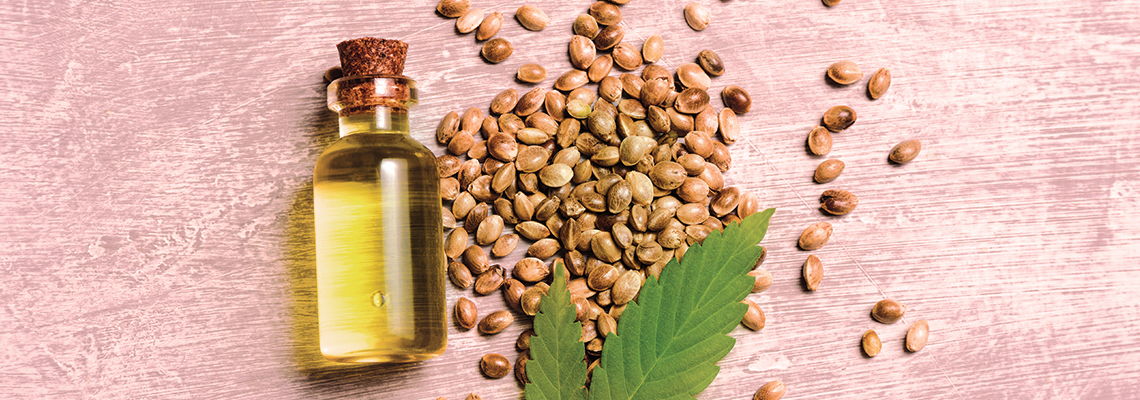 Glass bottle of oil with seeds and a green leaf on a pink background.