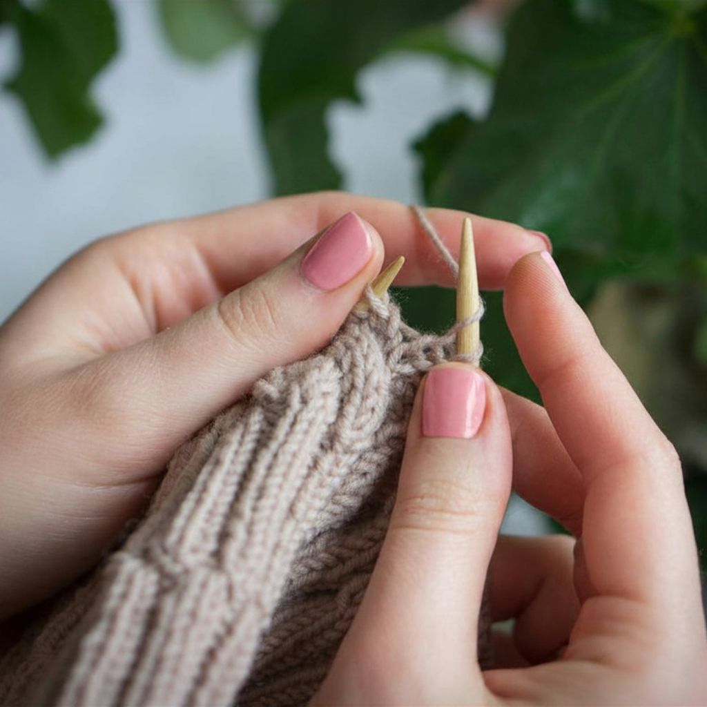 Person die mit einem beigen Garn und Holznadeln strickt.