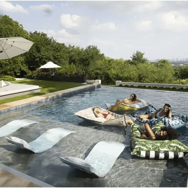 In-pool lounge chairs and floating pool seating in a low-maintenance modern backyard with umbrellas and mature landscaping