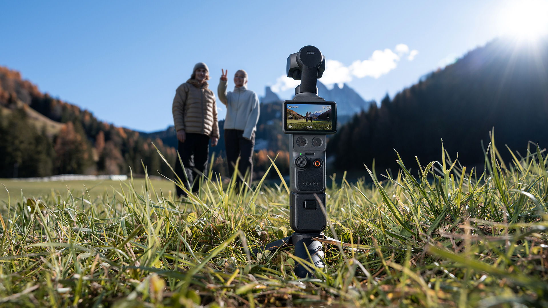 A DJI Osmo Pocket 4 on tripod with people in background and mountains. One of the people is making a V peace sign gesture with her hand.