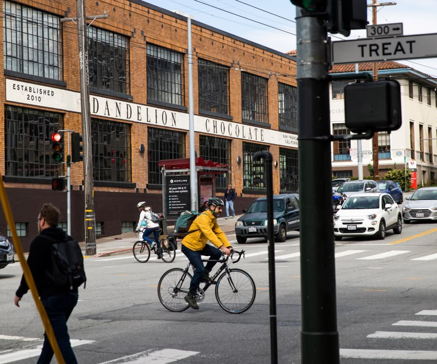 Bicyclist in yellow jacket crossing street near brick building and traffic signals.