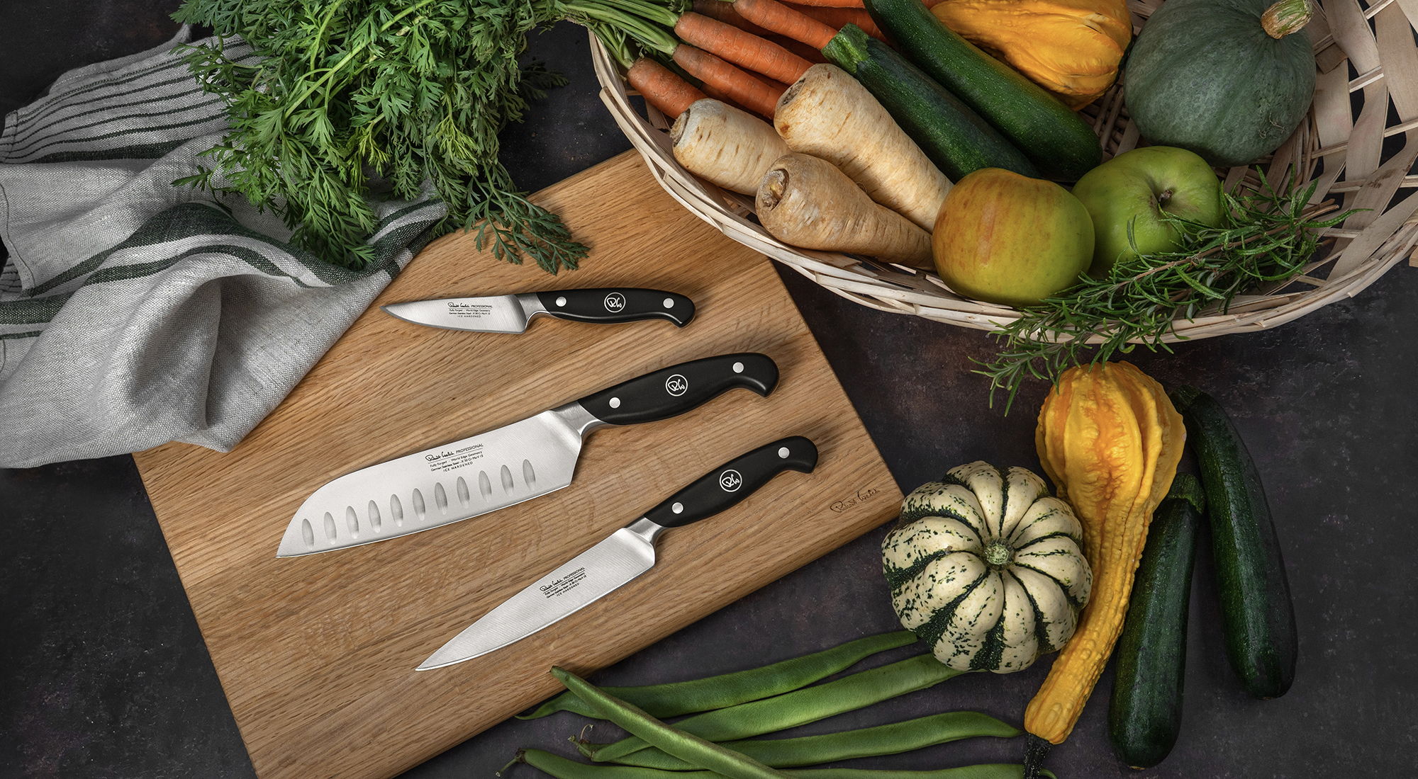 Professional knives on a cutting board with fresh vegetables and herbs.
