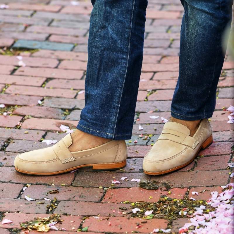 A close-up outdoor lifestyle shot from the shins down of a person standing on a red brick walkway scattered with pink cherry blossom petals. They are wearing dark wash denim jeans and a pair of light cream or desert suede Adelante Lorenzo penny loafers with a classic leather strap across the vamp and a thin leather sole.