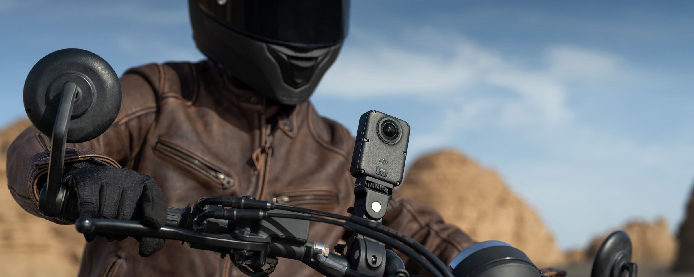 Person in a motorcycle helmet and jacket, riding a motorcycle in a desert landscape. A DJI Osmo 360 II is mounted to the motorcycle's handlebars.