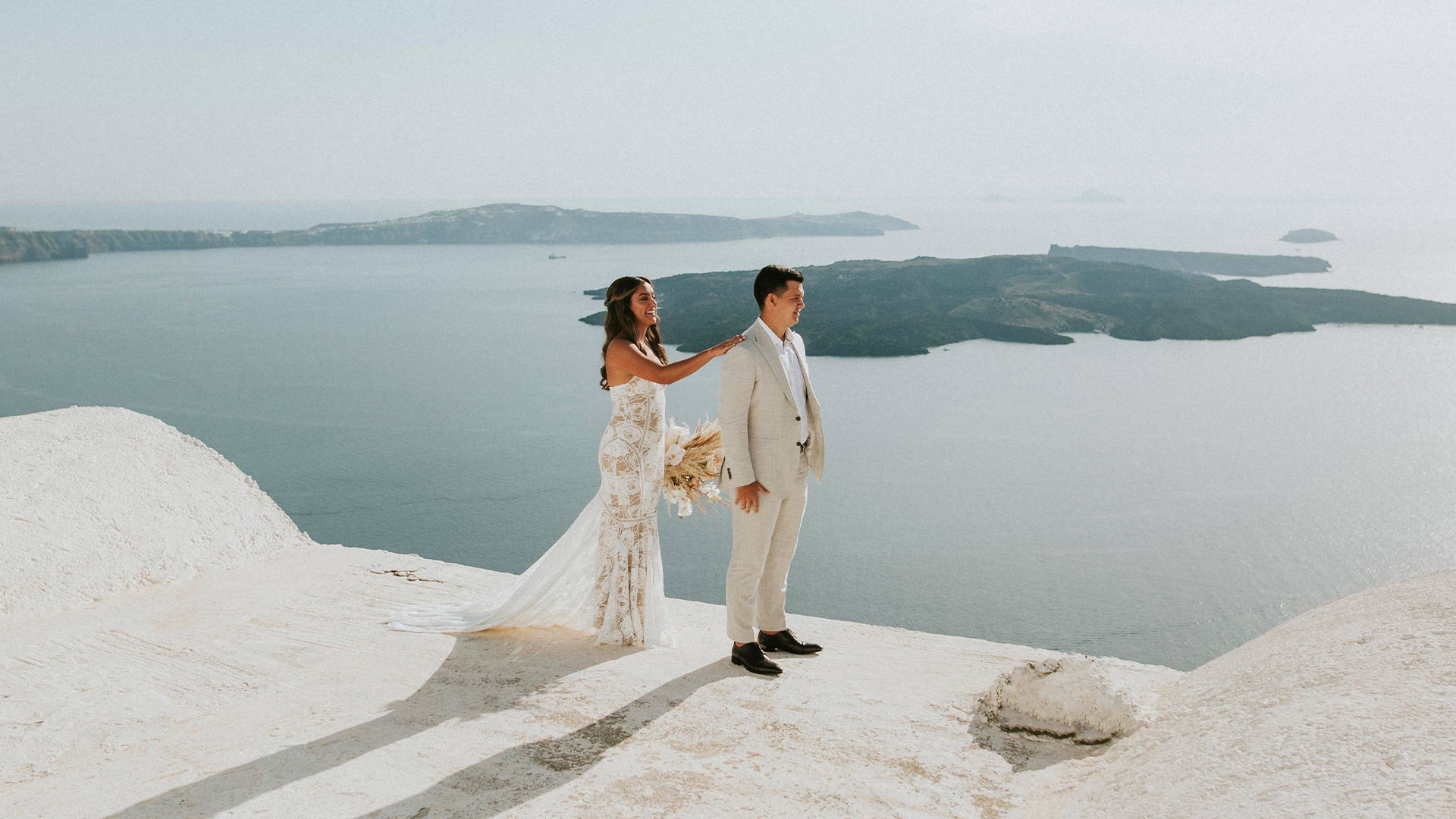 Mariée portant une robe de mariée en palmier à Santorin, en Grèce, tapant le marié sur l'épaule.