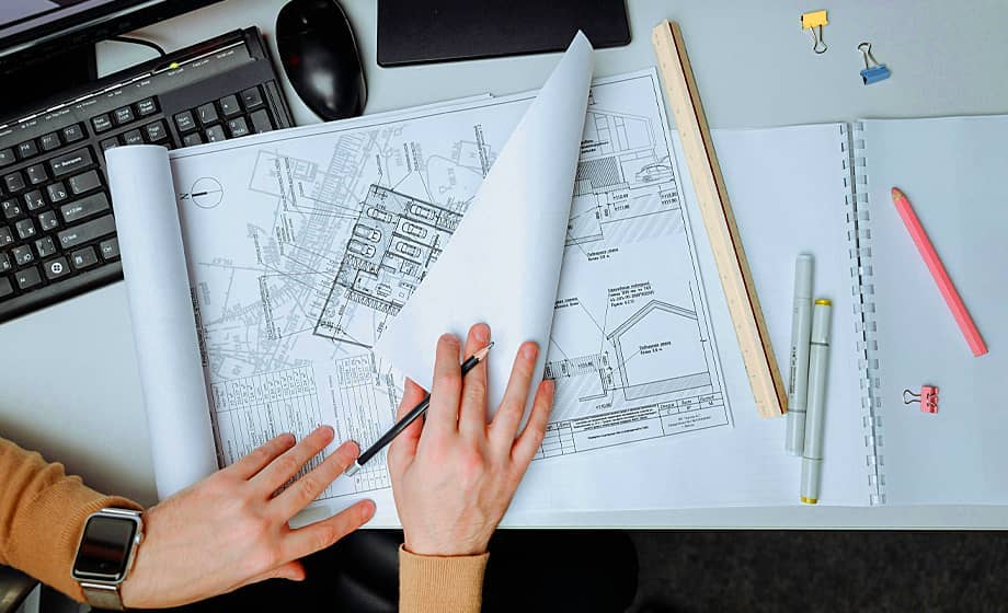 Hands reviewing architectural blueprints on a desk with stationery and a computer.