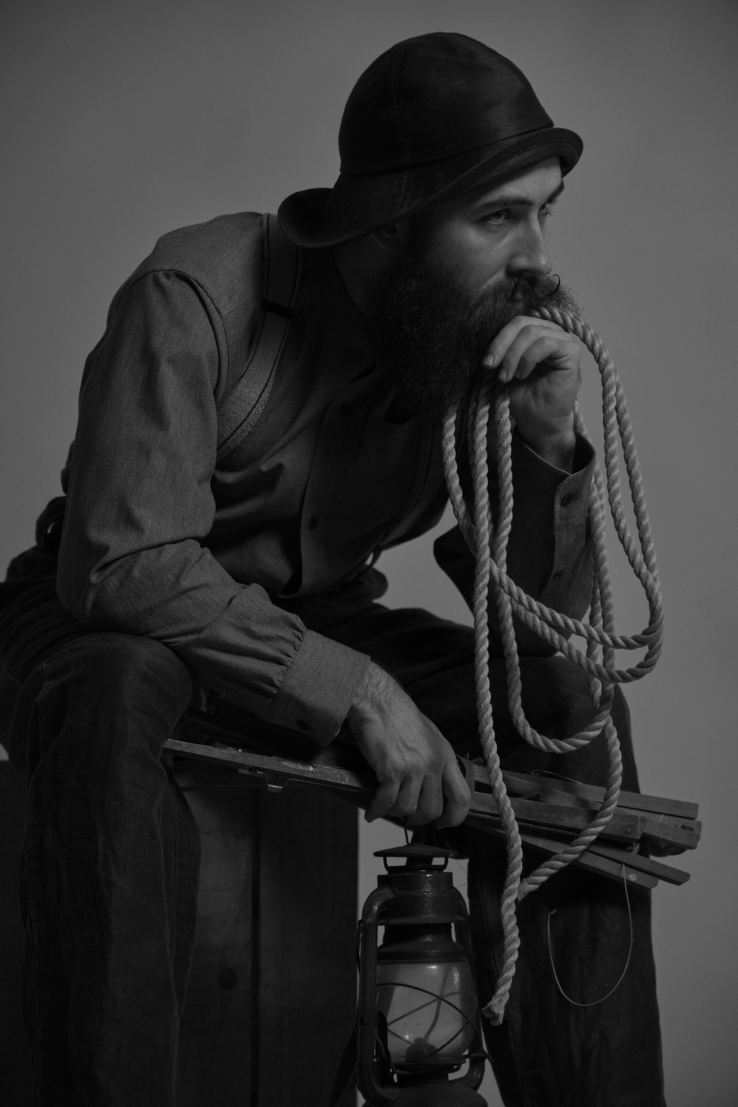 A man with a beard wearing suspenders, holding a lantern and rope.