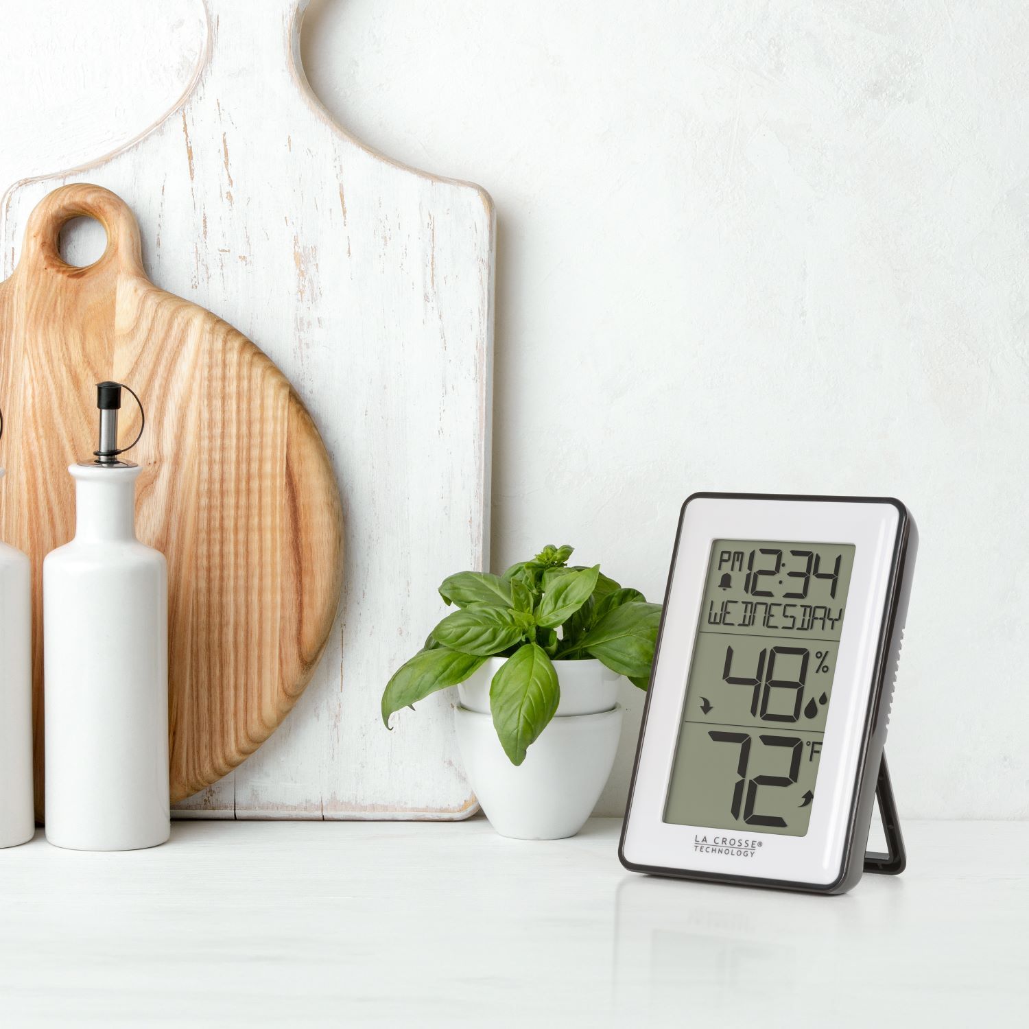 Digital thermometer displaying temperature and humidity next to a plant and kitchen items.