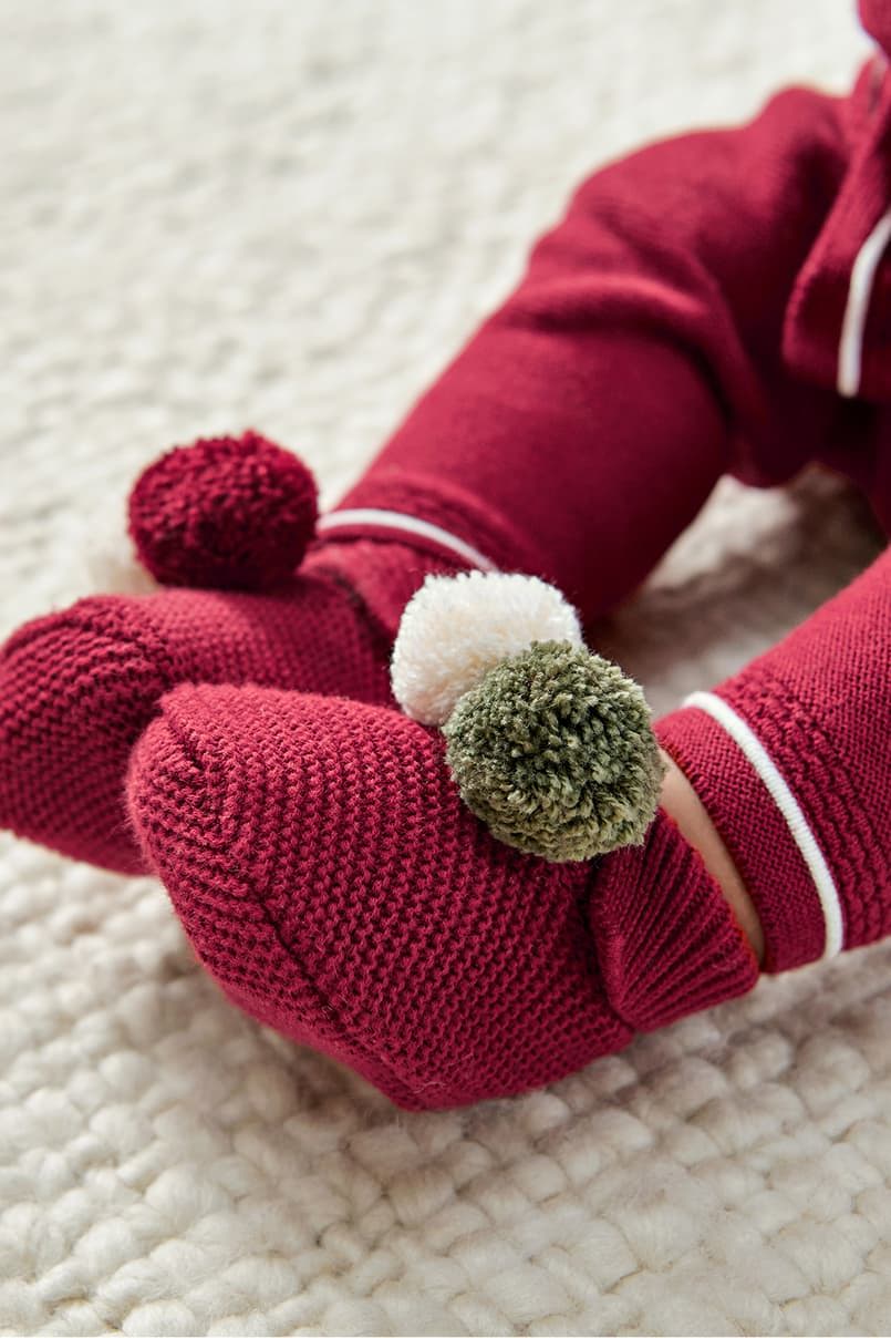 Red mittens with colorful pom-poms on a textured surface.