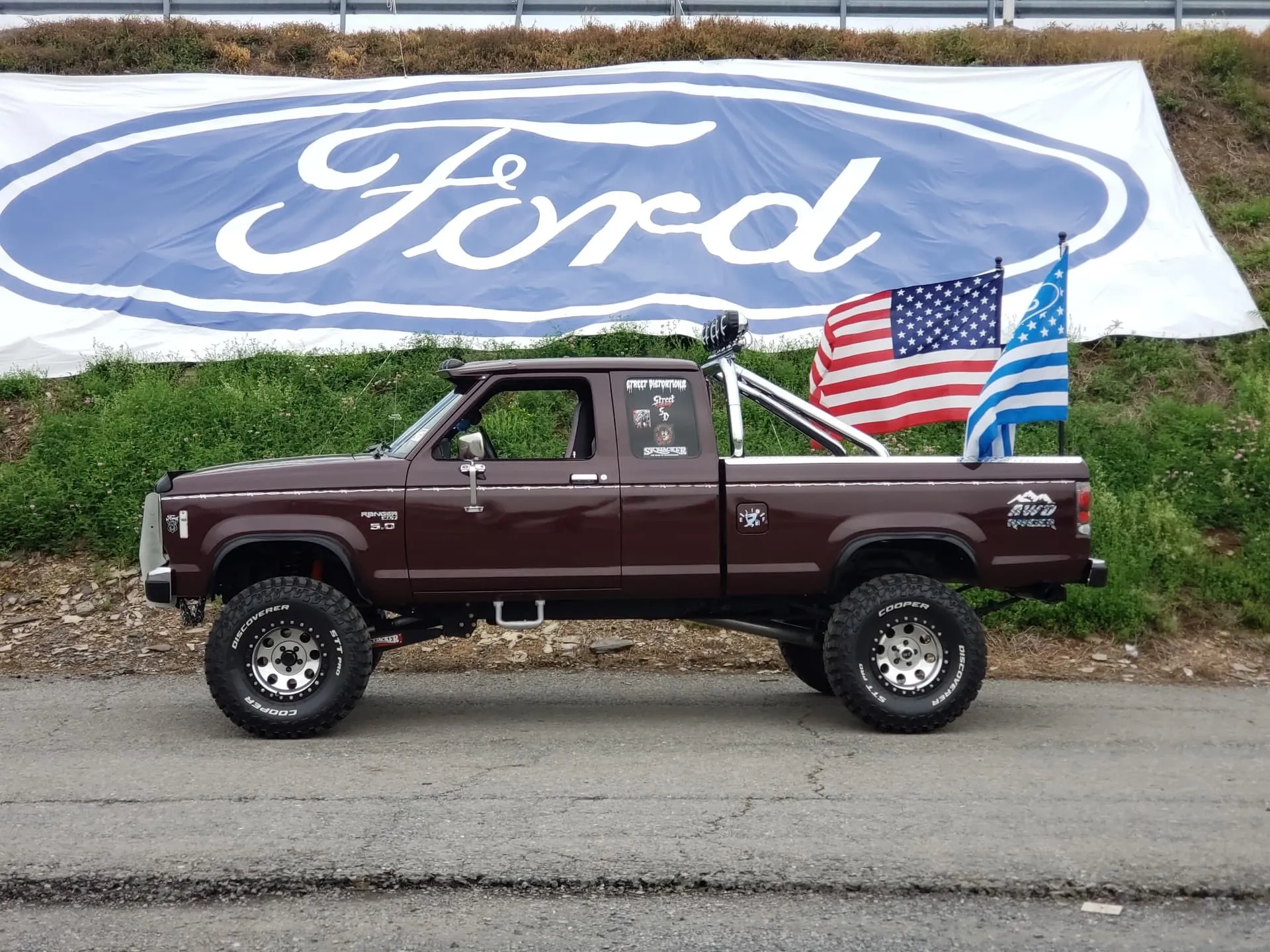 Side profile Brenda's lifted maroon 1988 Ford Ranger with roll bar and flags in front of Ford banner photo 2