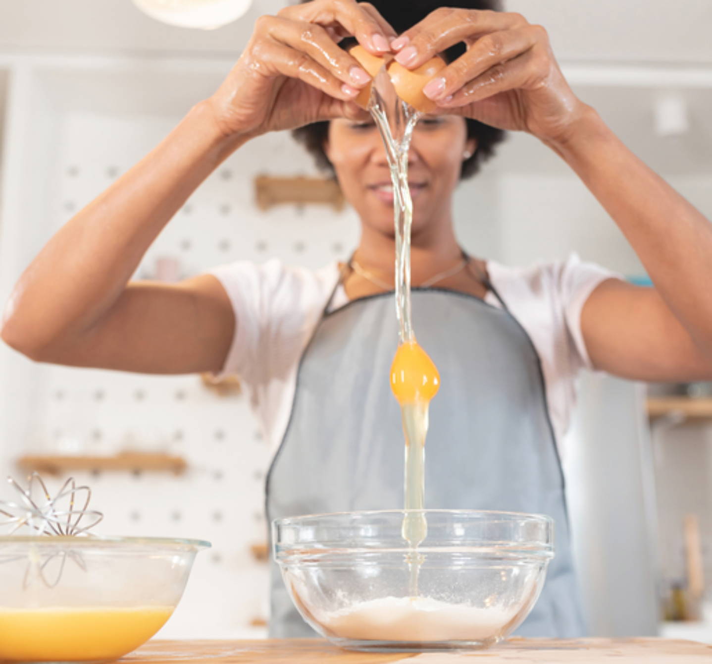 Une femme casse un œuf en hauteur au-dessus d’un bol, de sorte que le blanc et le jaune tombent en filet dans la farine - l’œuf peut provoquer des allergies.