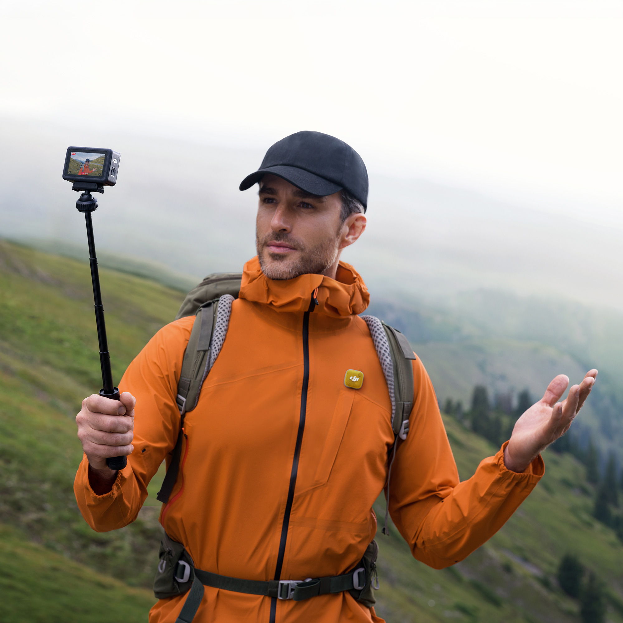 Person in orange jacket holding a Osmo Action on a selfie stick and recording audio with DJI Mic Mini 2 in a scenic landscape.