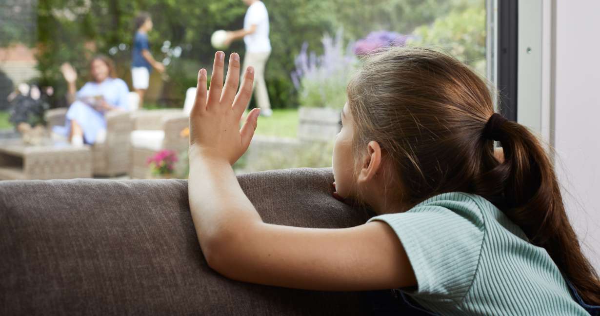 Petite fille qui touche la fenêtre en regardant tristement sa famille jouer dans le jardin; son rhume des foins l’oblige parfois à rester à l’intérieur.
