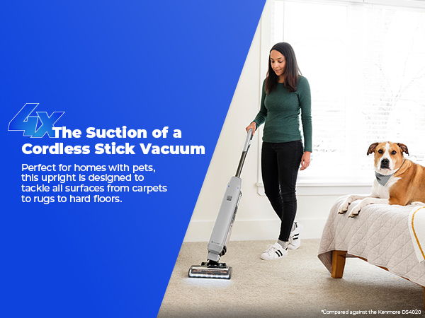 A woman using a cordless stick vacuum on carpet near a dog on a bed.