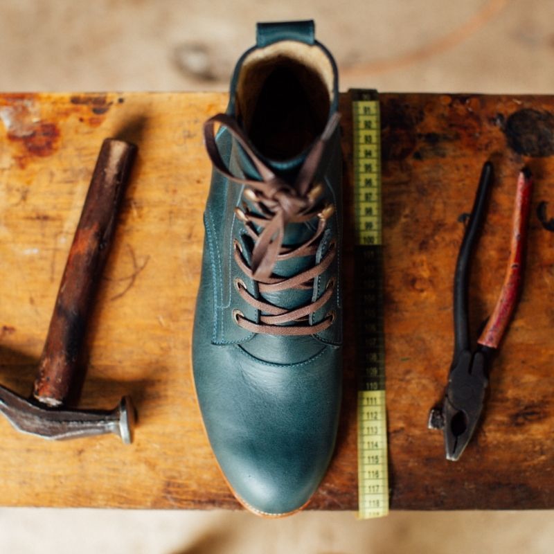 A top-down overhead shot of a single denim blue leather Adelante Havana boot sitting vertically on a rustic wooden cobbler's workbench. The lace-up boot features dark brown laces and is neatly surrounded by traditional shoemaking tools, including a worn wooden-handled cobbler's hammer on the left, a yellow measuring tape alongside the boot, and a pair of vintage pliers on the right.