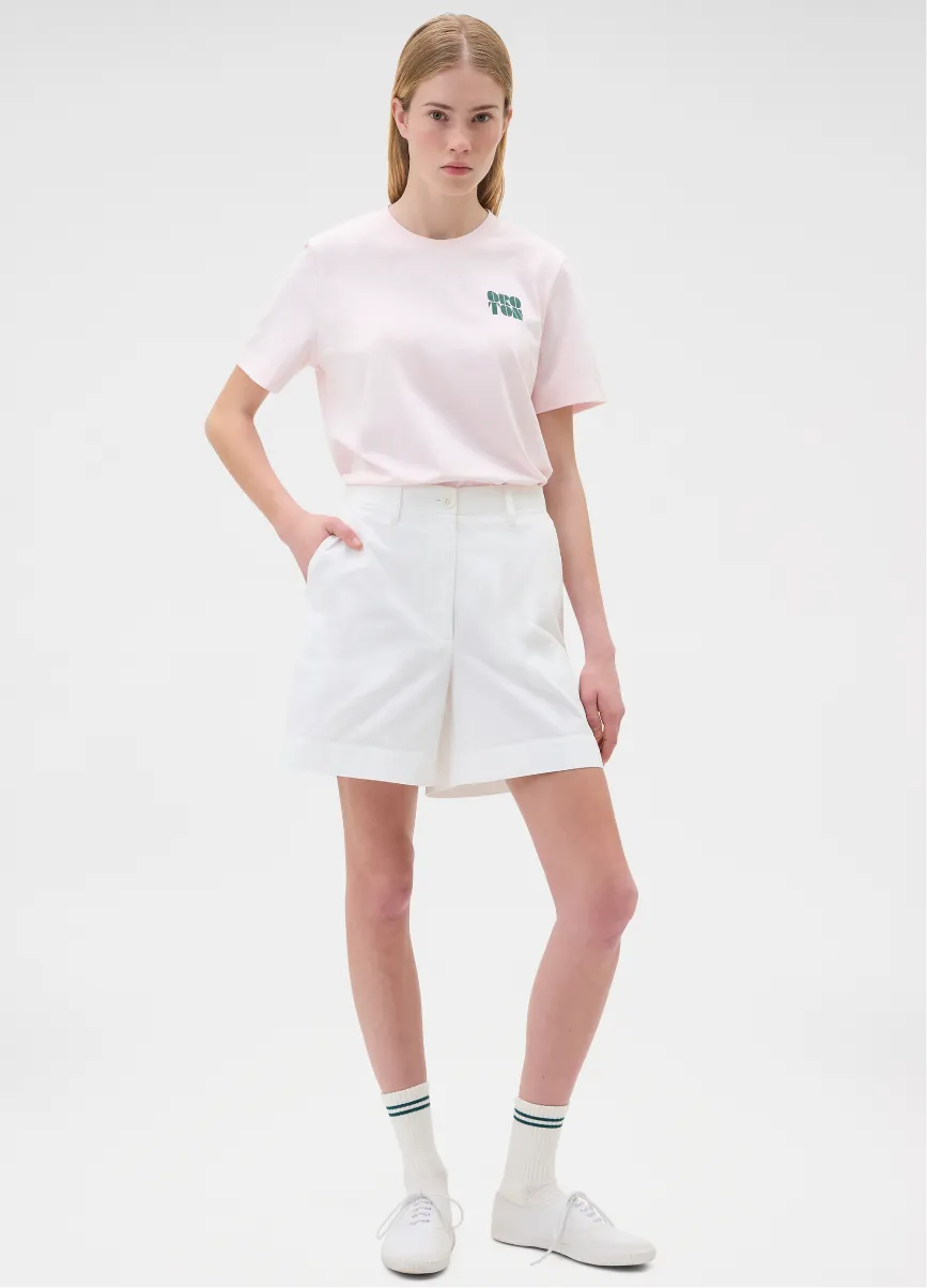 A woman in a pink shirt and white shorts stands against a plain background.