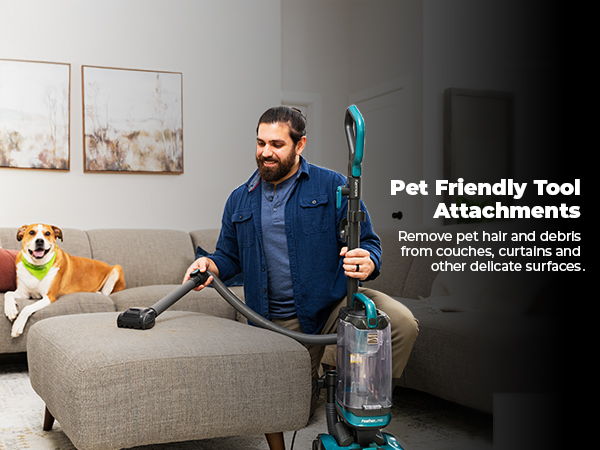 Man vacuuming with pet-friendly tool, dog sitting on couch nearby.