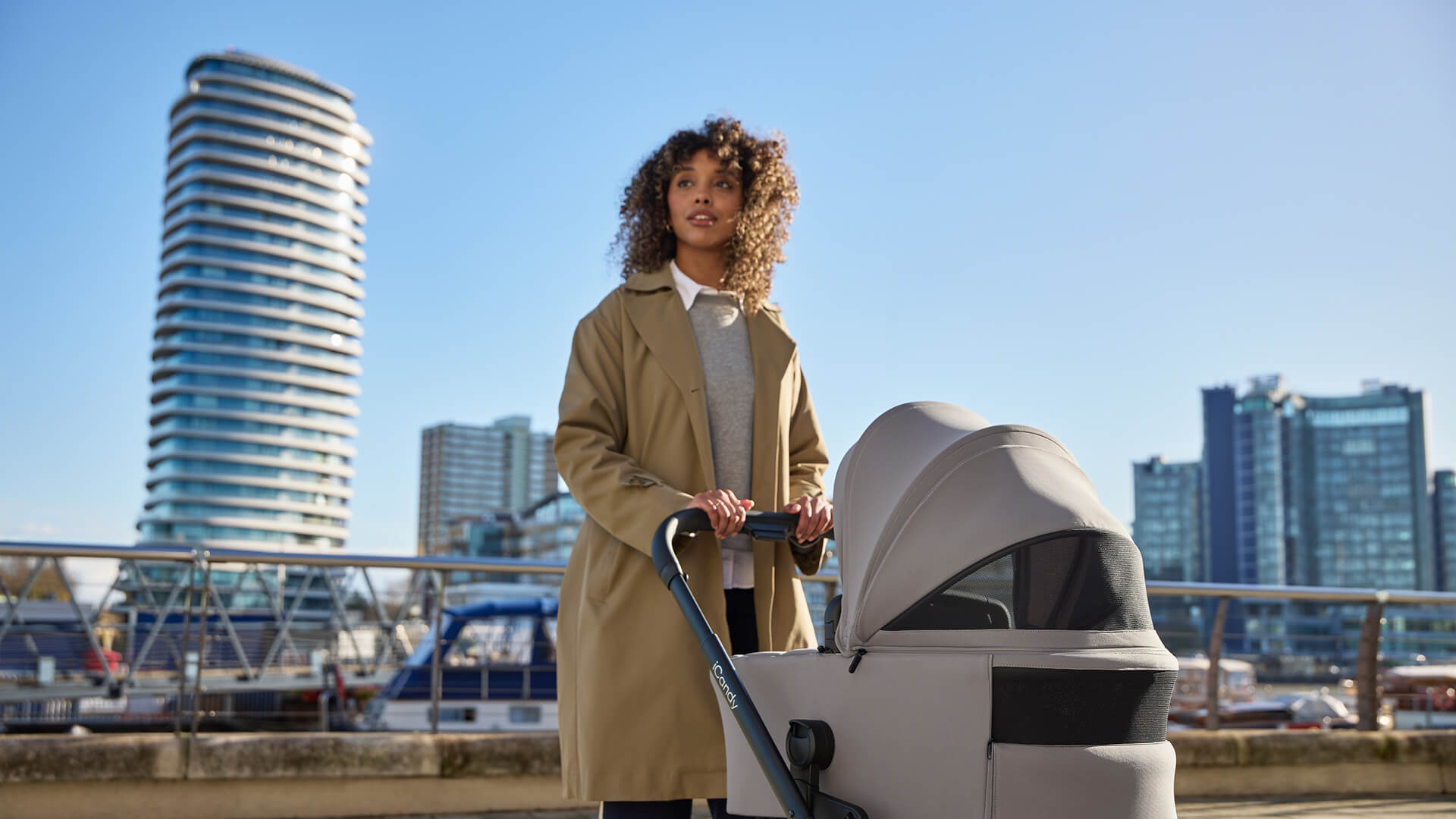 Woman with curly hair pushing a iCandy stroller near a city skyline.