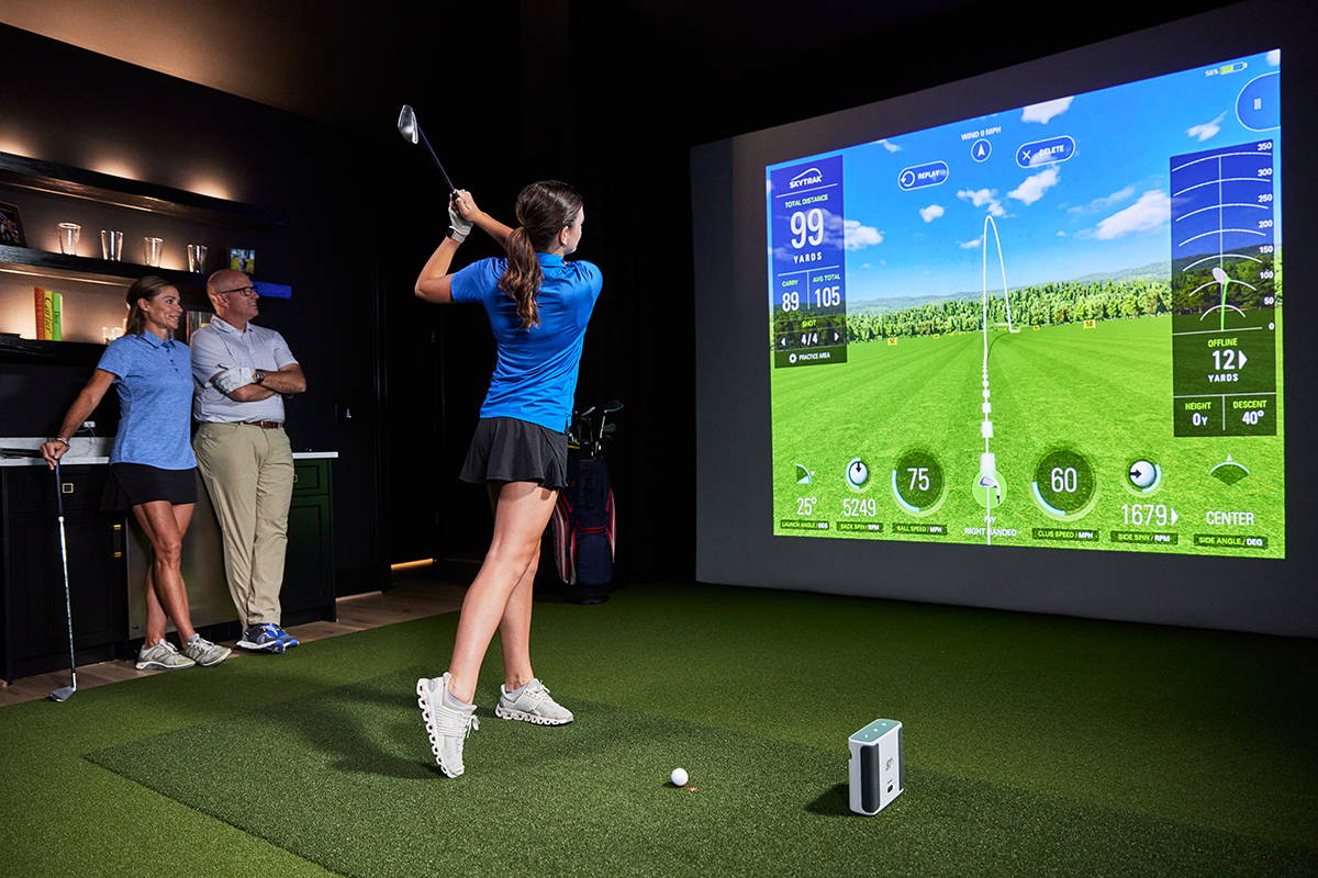 A girl swings in a SkyTrak+ golf simulator with impact screen while a man and woman watch