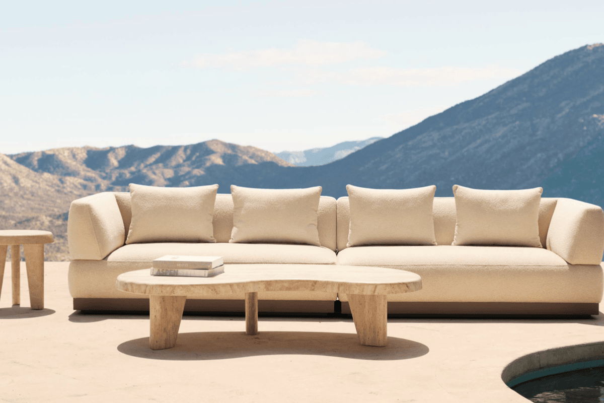 Cream outdoor sofa with matching throw pillows behind a travertine coffee table on a sunlit terrace with mountain views and a pool edge.