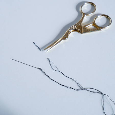  A double threaded needle showing a knot at the very end with its trimmed tail on a white table with golden stork scissors