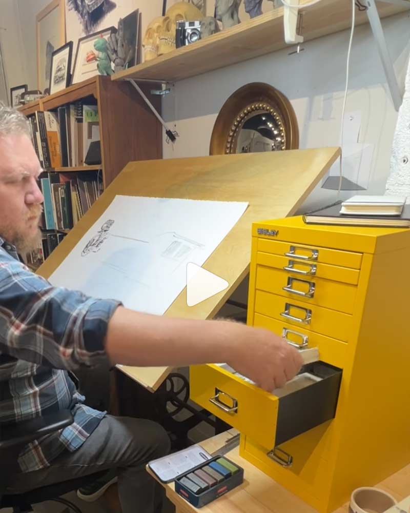 Person working at a desk with a yellow filing cabinet and drawing materials.