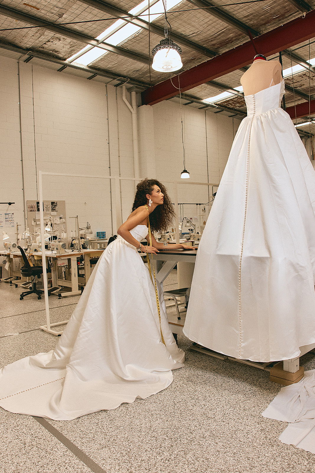 A woman in a white dress examines a larger white gown in a workshop.