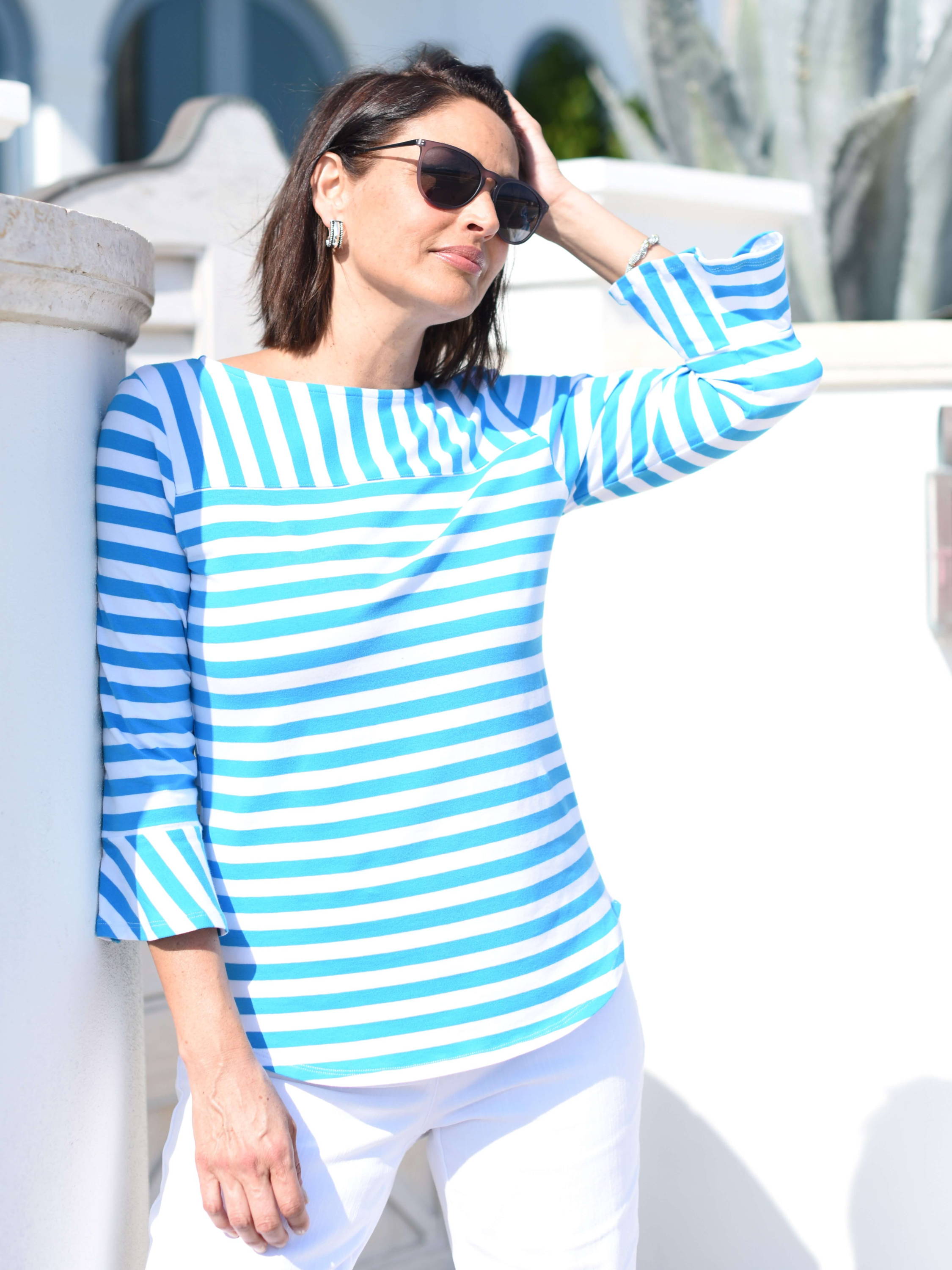 A woman in a blue and white striped top  and sunglasses leans against a white wall.