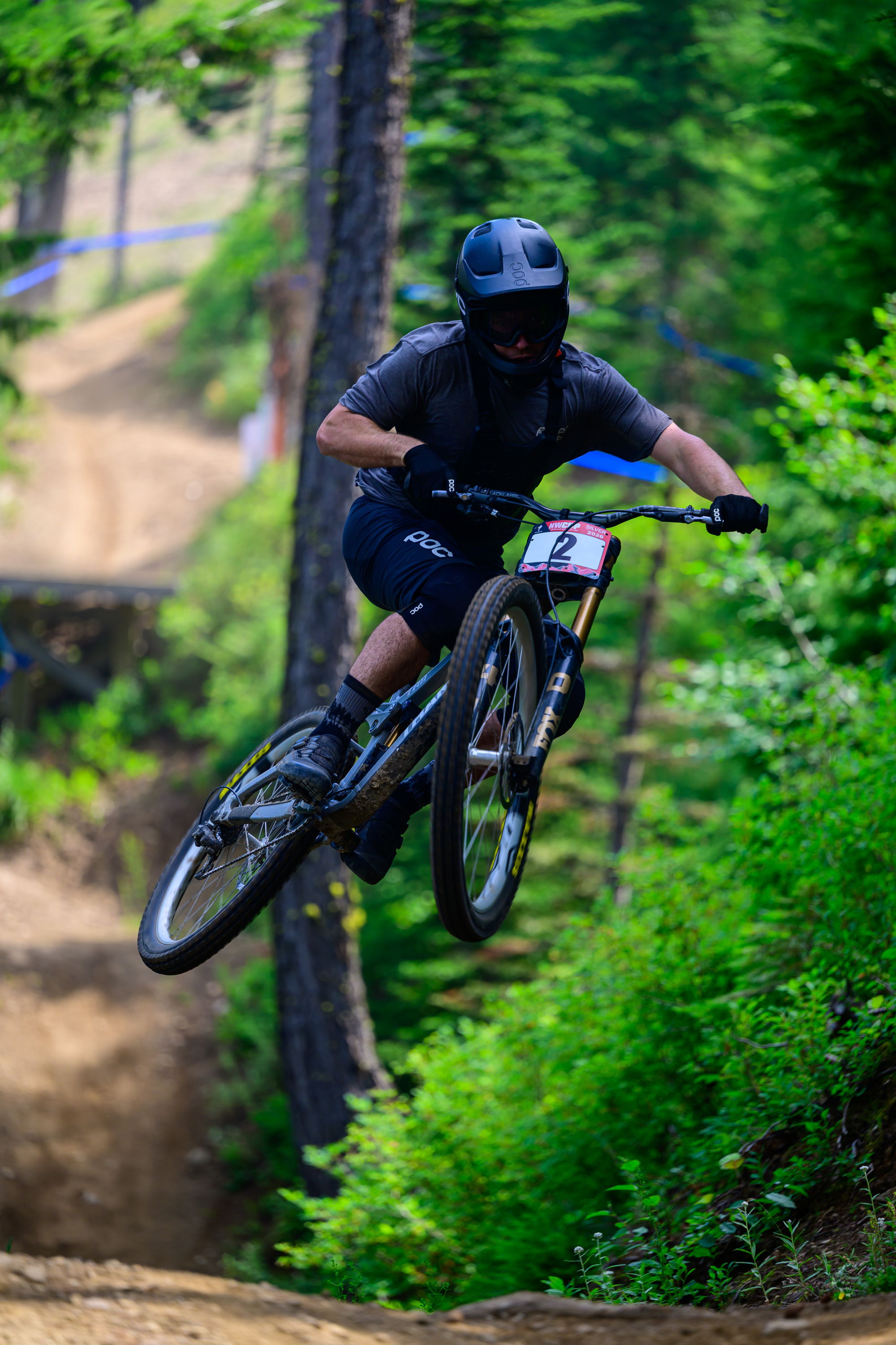 Mountain biker performing a jump on a dirt trail surrounded by trees.