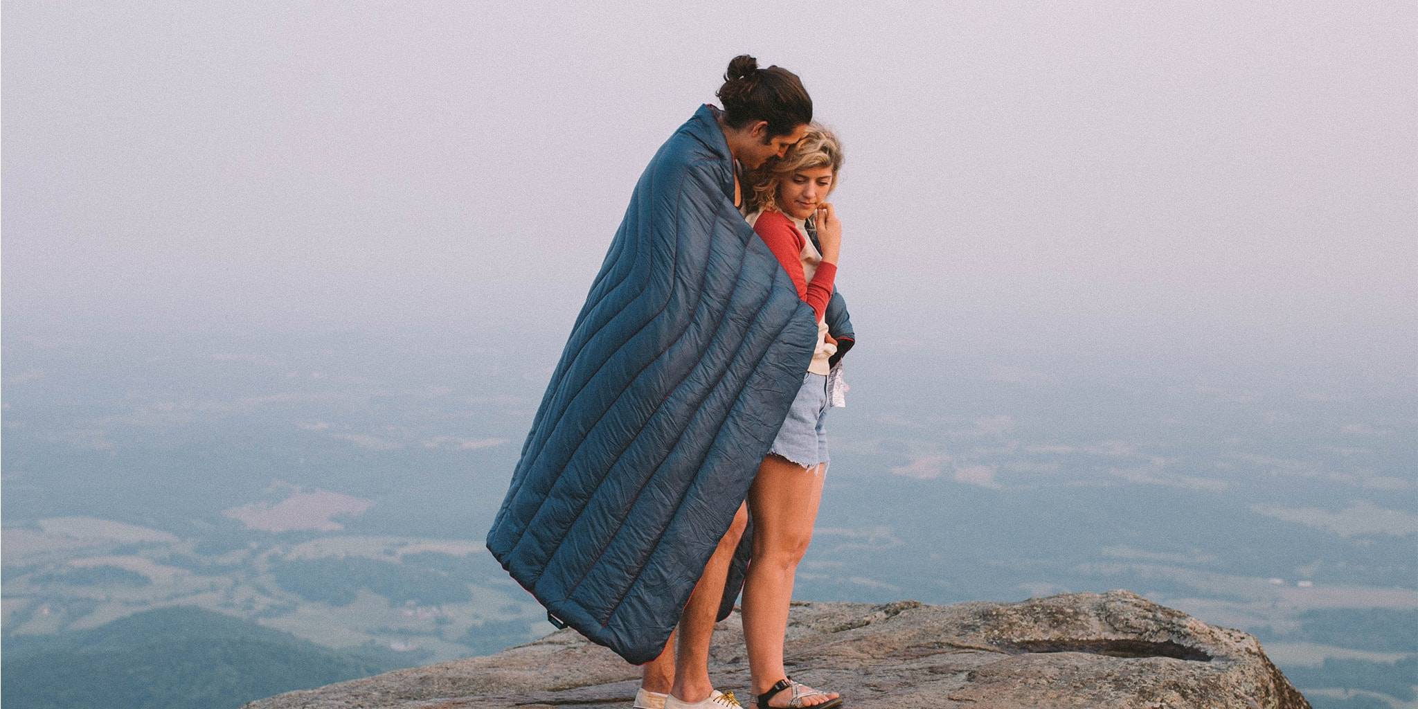 Man holding a woman on a rock cliff wearing a blue Rumpl blanket