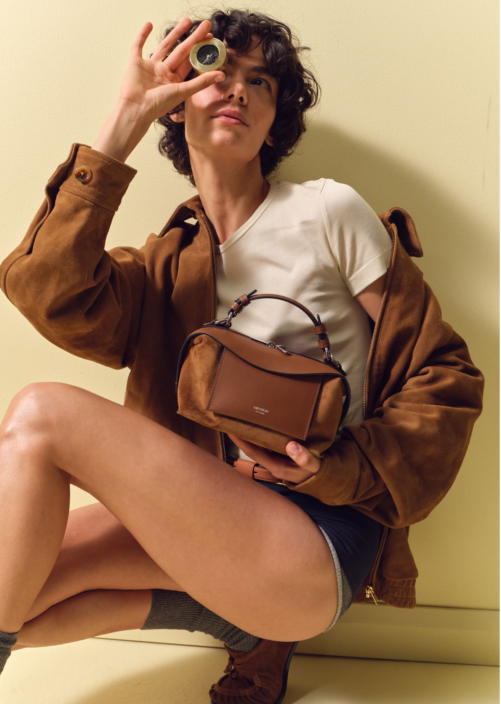 Person in stylish outfit holding a bag and looking through a lens.