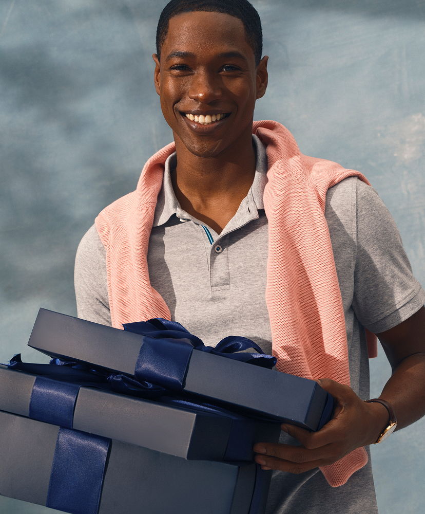 Smiling young man holding two gift boxes, wearing a pink sweater over his shoulders.