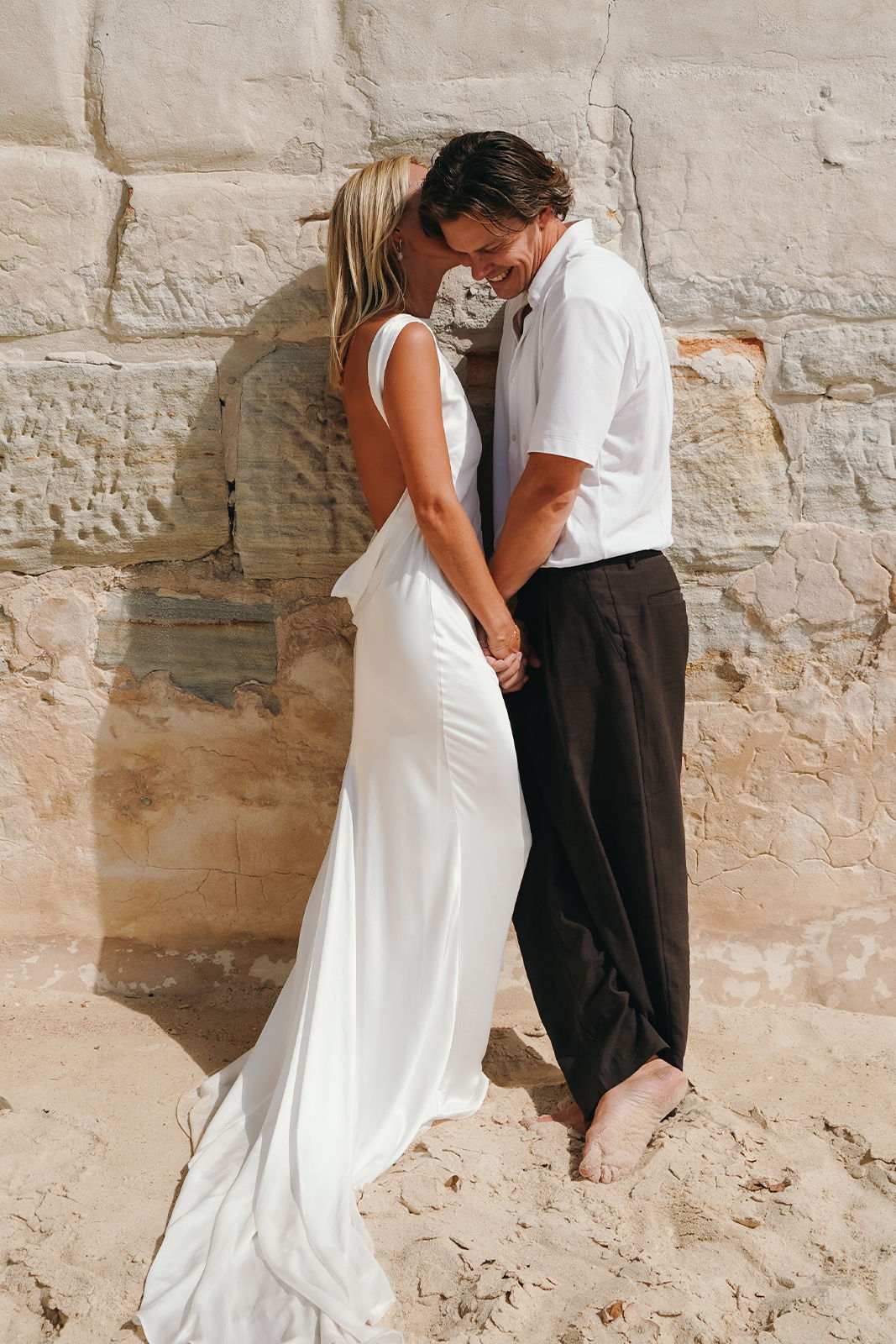 Couple in wedding attire embracing against a stone wall at the beach.
